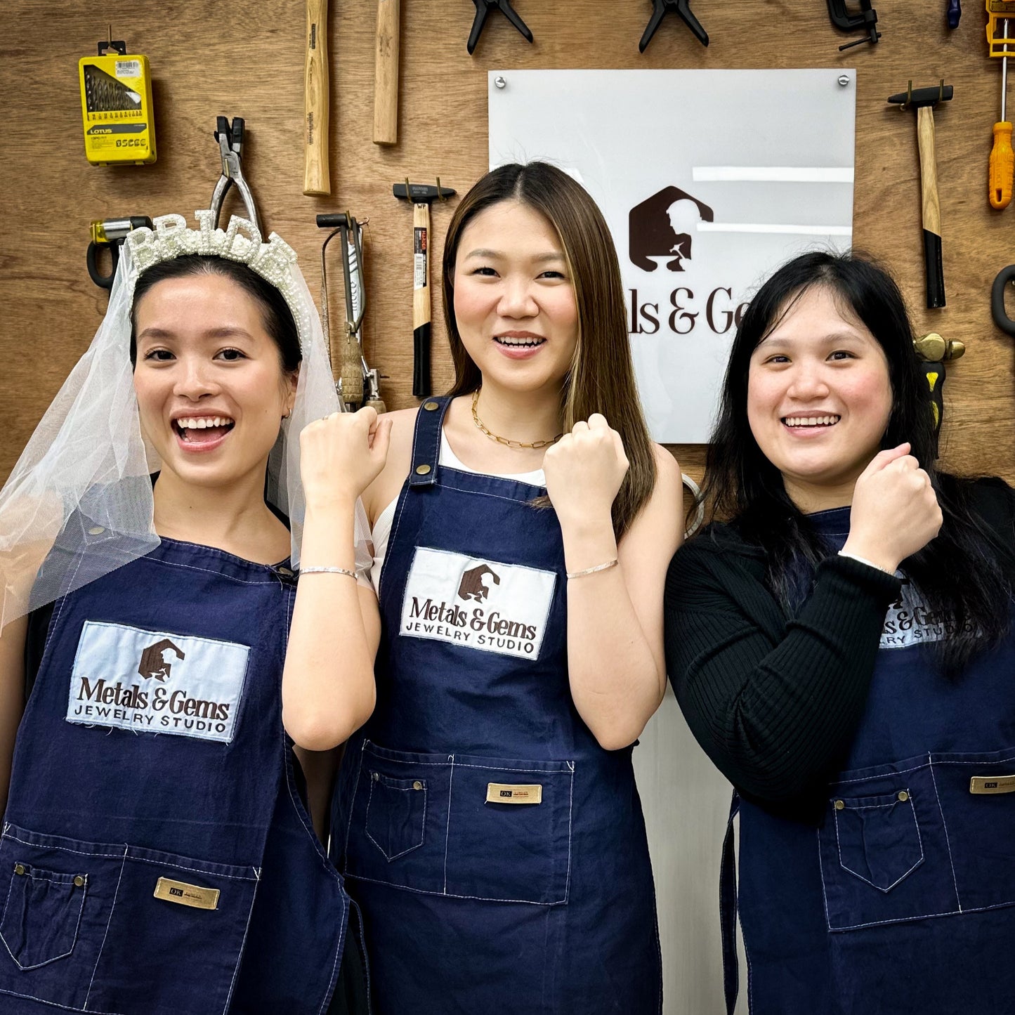 Bridal shower jewelry making three women wearing blue aprons with a logo, posing in front of a wall with tools and equipment.