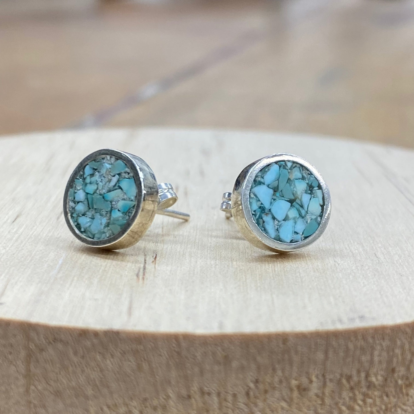 A pair of round stud earrings with light blue marbled resin centers and silver-toned bezels, displayed on a light wood slice against a blurred wooden background.