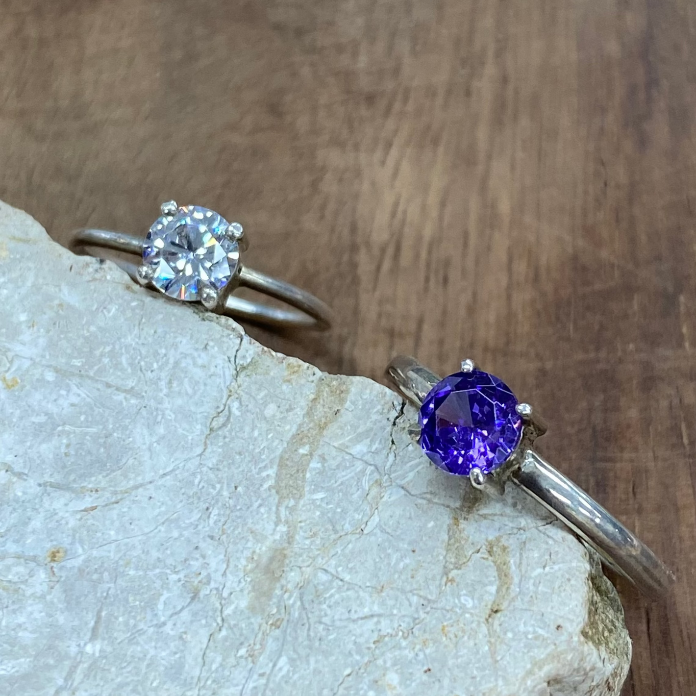 Two silver rings displayed on a light stone slab over a wooden surface. One ring features a round clear gemstone, and the other features a round purple gemstone.