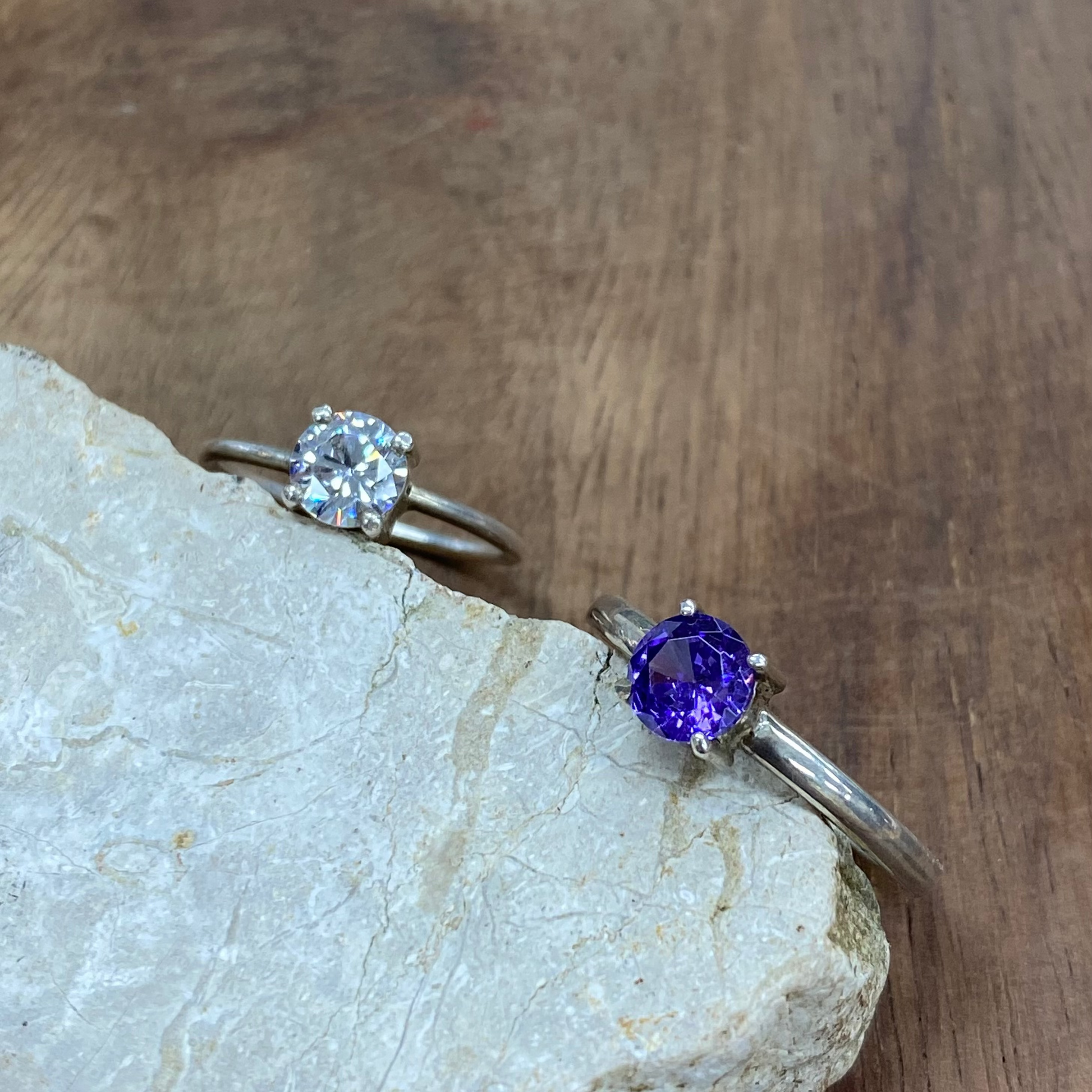 Two silver rings displayed on a light stone slab over a wooden surface. One ring features a round clear gemstone, and the other features a round purple gemstone.