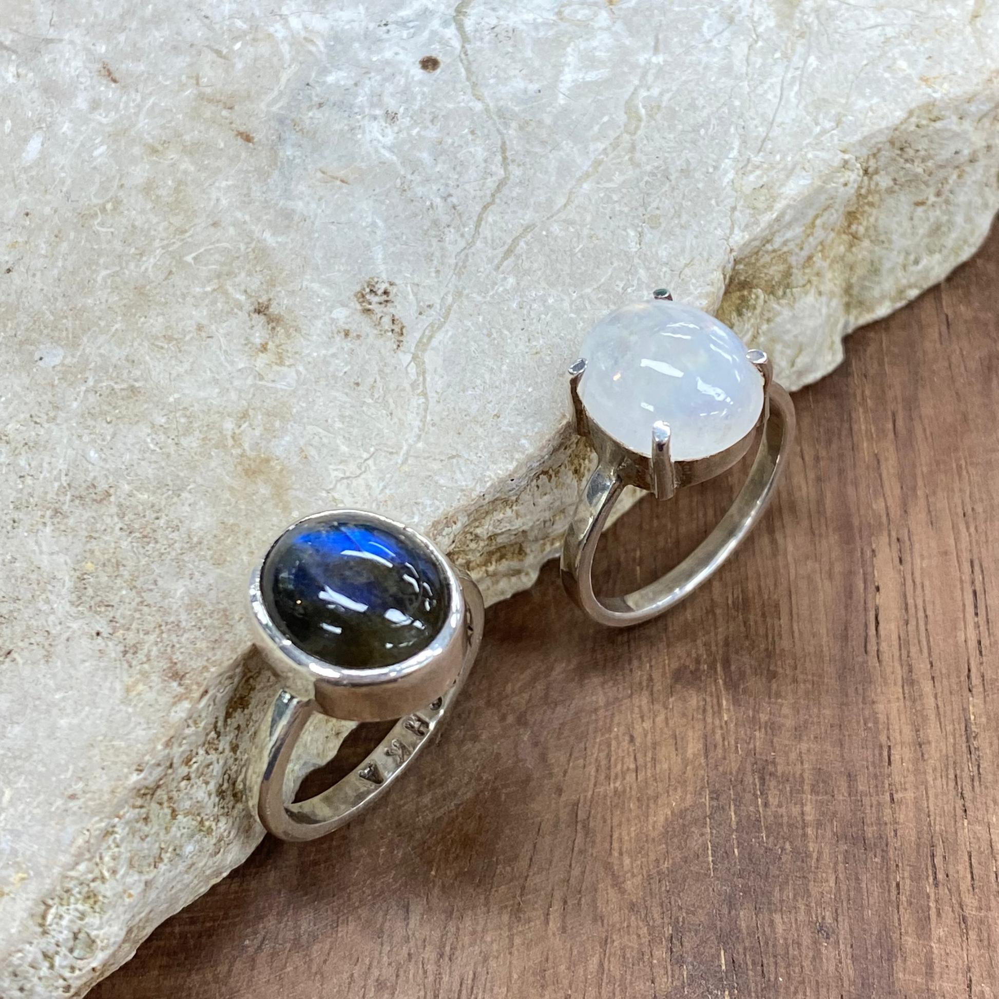 Two sterling silver rings displayed on a natural stone slab over a wooden surface. One ring features a dark, polished labradorite cabochon showing blue flashes, and the other features a milky white moonstone cabochon.
