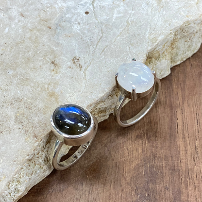 Two sterling silver rings displayed on a natural stone slab over a wooden surface. One ring features a dark, polished labradorite cabochon showing blue flashes, and the other features a milky white moonstone cabochon.