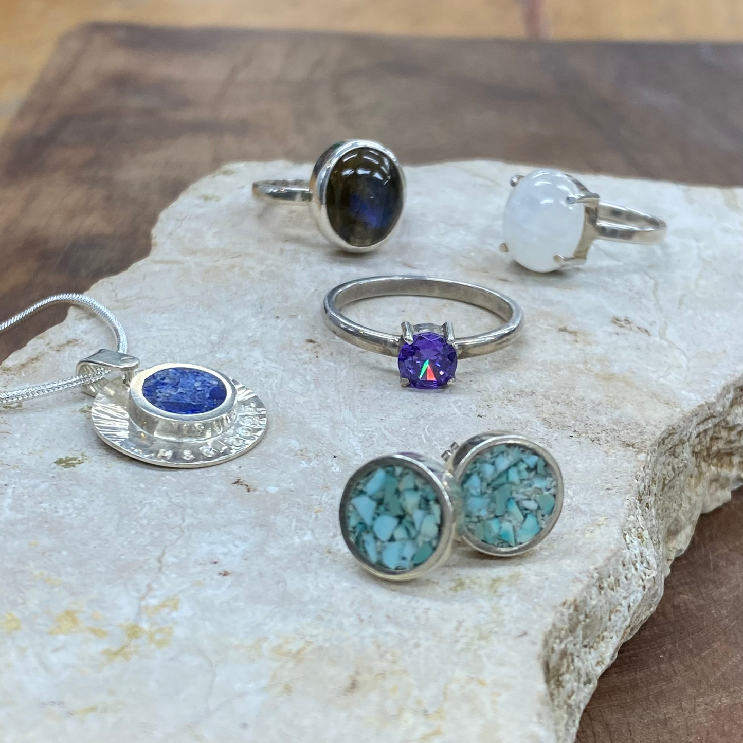 A collection of silver jewelry including rings, stud earrings, and a pendant, displayed on a natural stone slab over a wooden surface. The jewelry features various colored stones, including turquoise, lapis lazuli, a purple topaz, and a white moonstone.