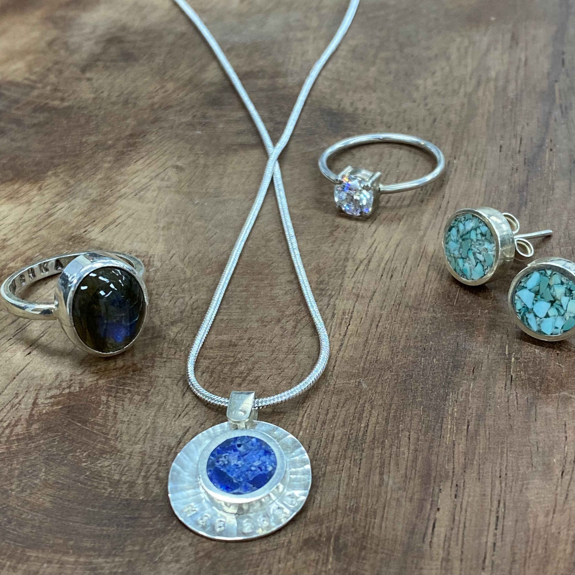 A jewelry set displayed on a rustic wooden surface, featuring a silver pendant necklace with a round blue stone, a silver ring with a dark oval stone, a silver band with a clear stone, and a pair of silver stud earrings with turquoise-blue marbled stones.
