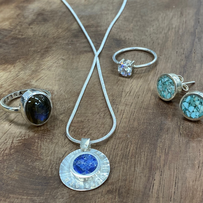 A jewelry set displayed on a rustic wooden surface, featuring a silver pendant necklace with a round blue stone, a silver ring with a dark oval stone, a silver band with a clear stone, and a pair of silver stud earrings with turquoise-blue marbled stones.