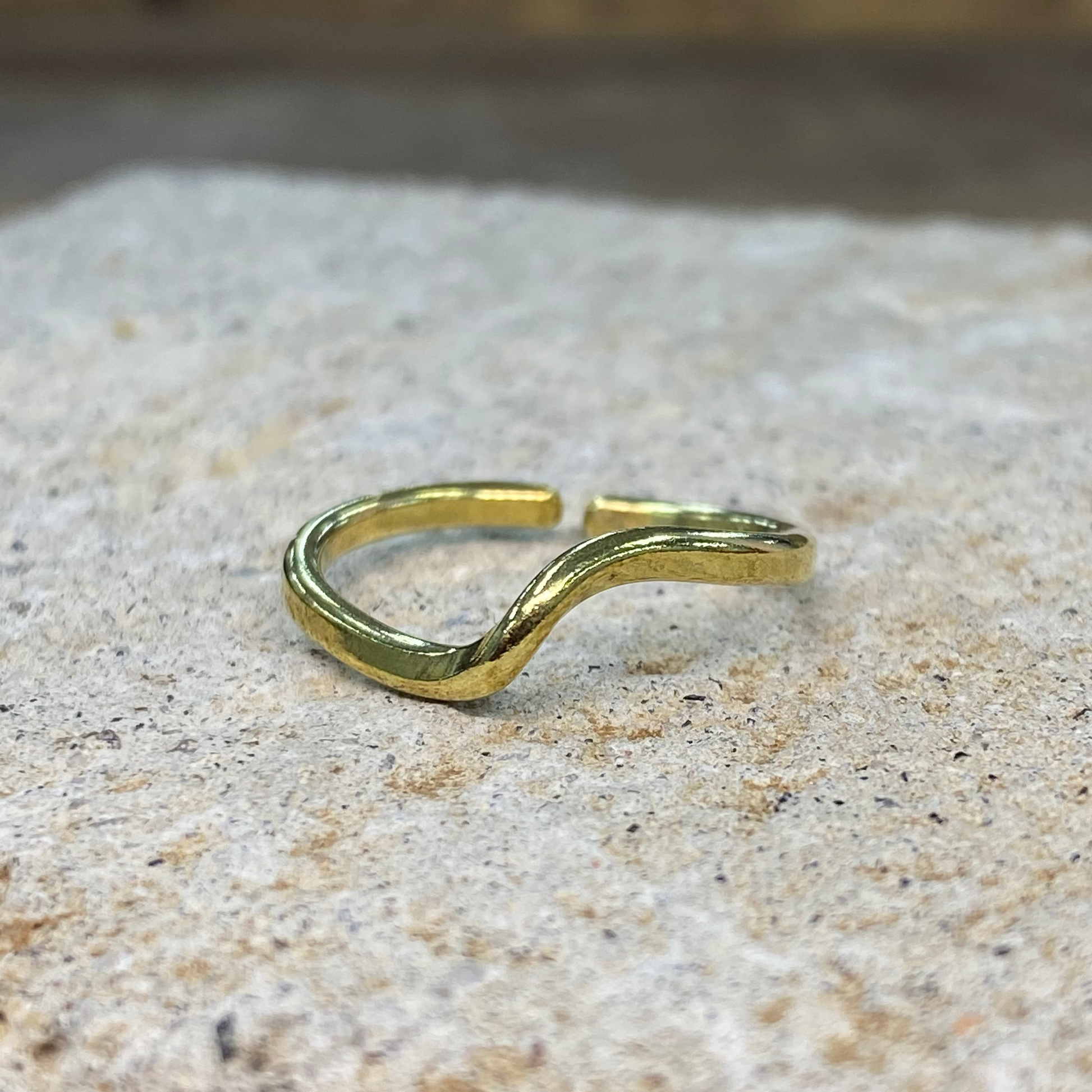 A single open cuff ring in polished yellow gold, resting on a textured light beige stone surface with a blurred neutral background.