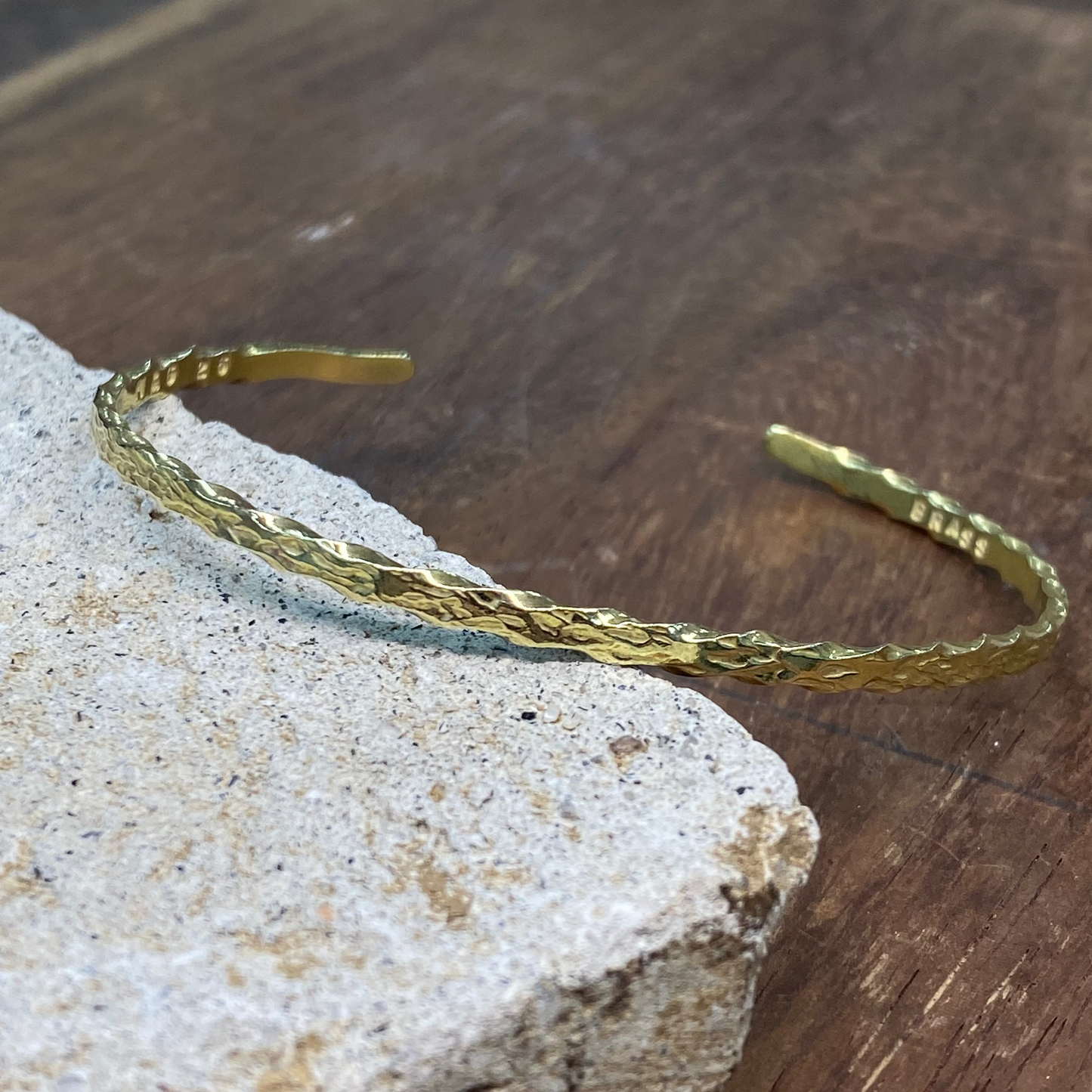 A textured gold-tone open cuff bracelet resting on a light gray stone over a dark brown wooden surface.