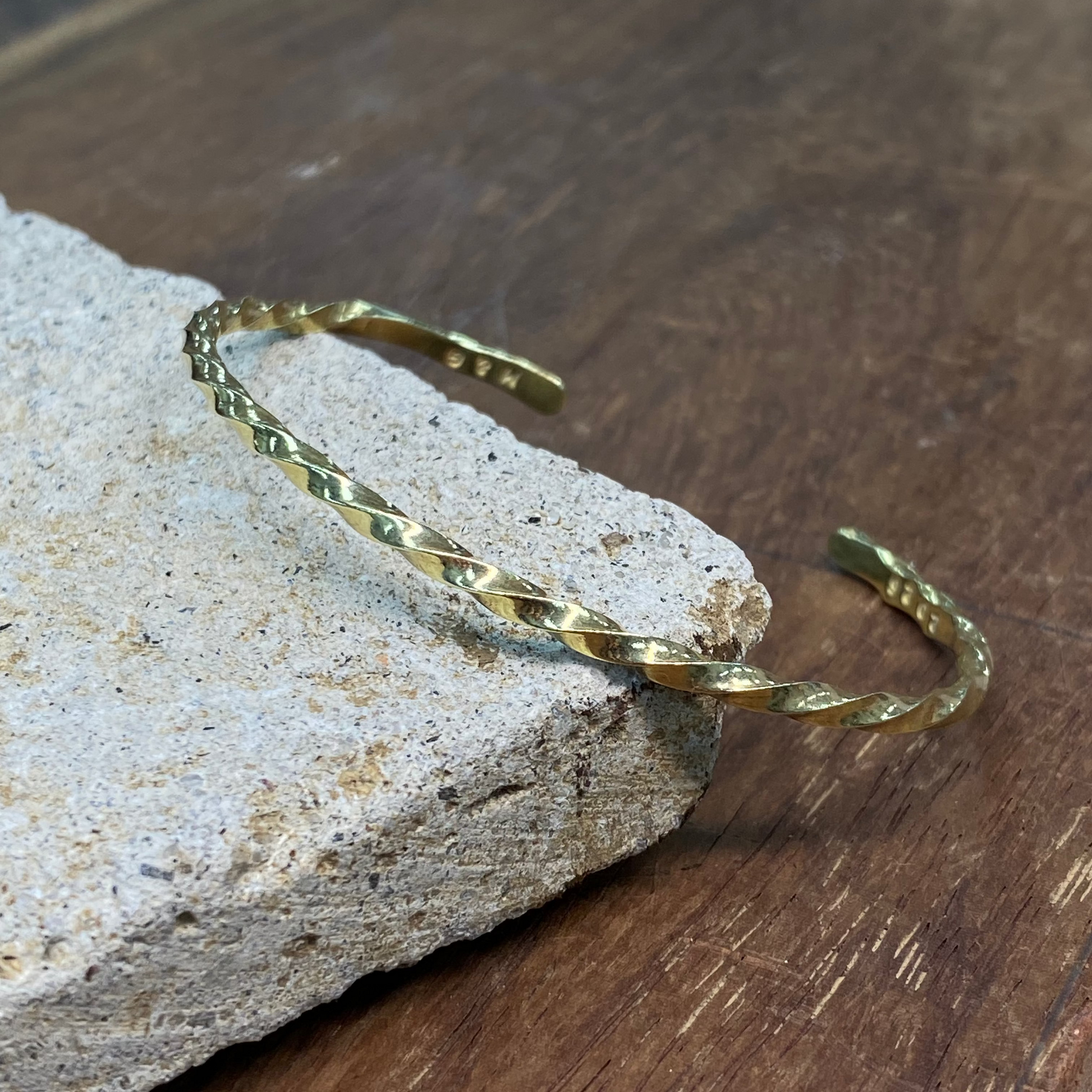 A gold-toned twisted open cuff bracelet resting on a textured light gray stone slab over a dark brown wooden surface.