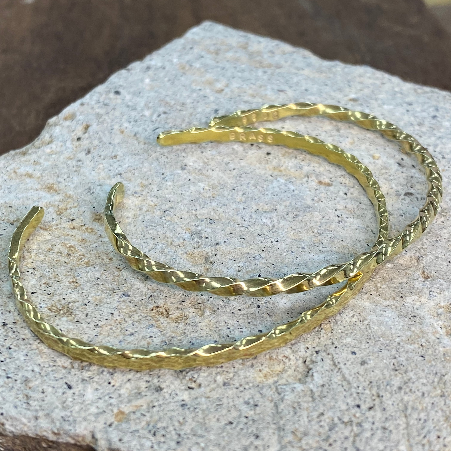 Two gold-toned twisted bangle bracelets resting on a textured light gray stone slab with a dark brown background.
