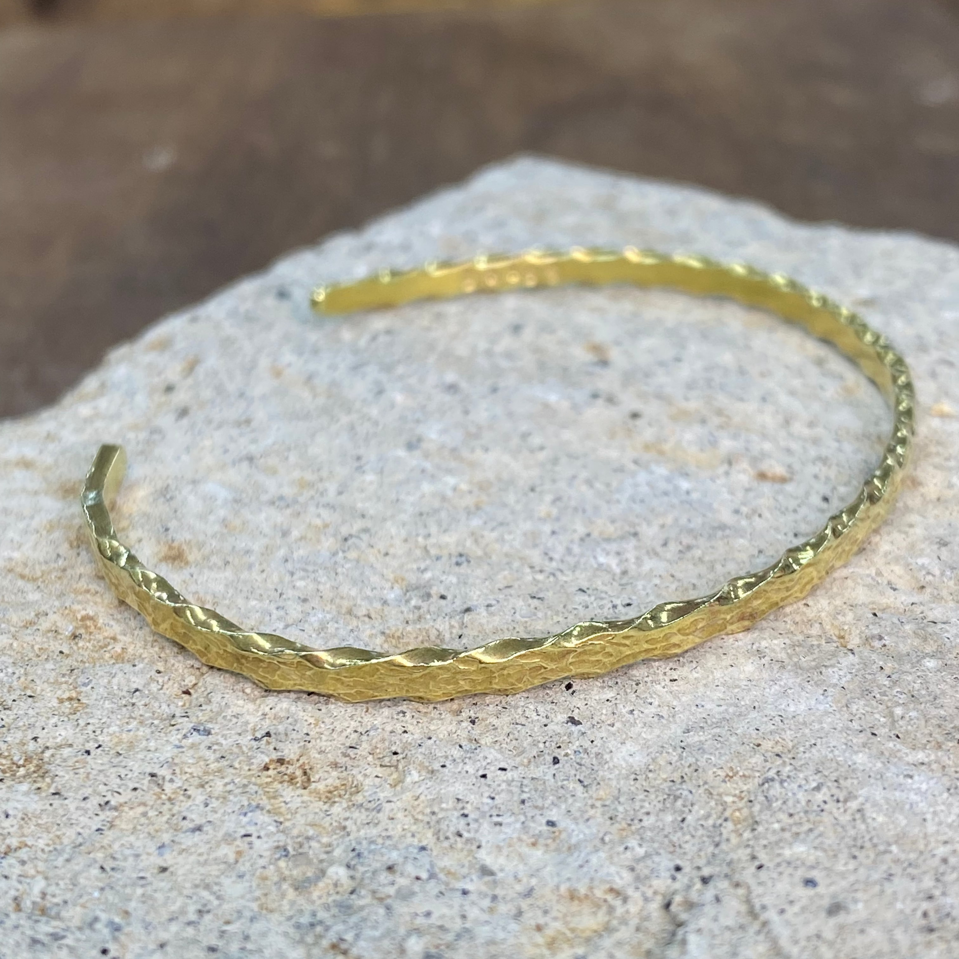 A textured gold-tone metal cuff bracelet resting on a light gray stone surface with a blurred brown background.