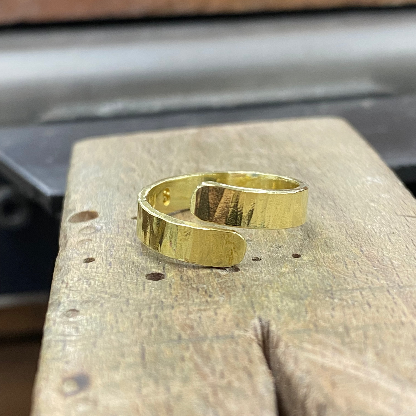 A polished gold-tone metal cuff ring with a hammered texture, resting on a rustic wooden block.