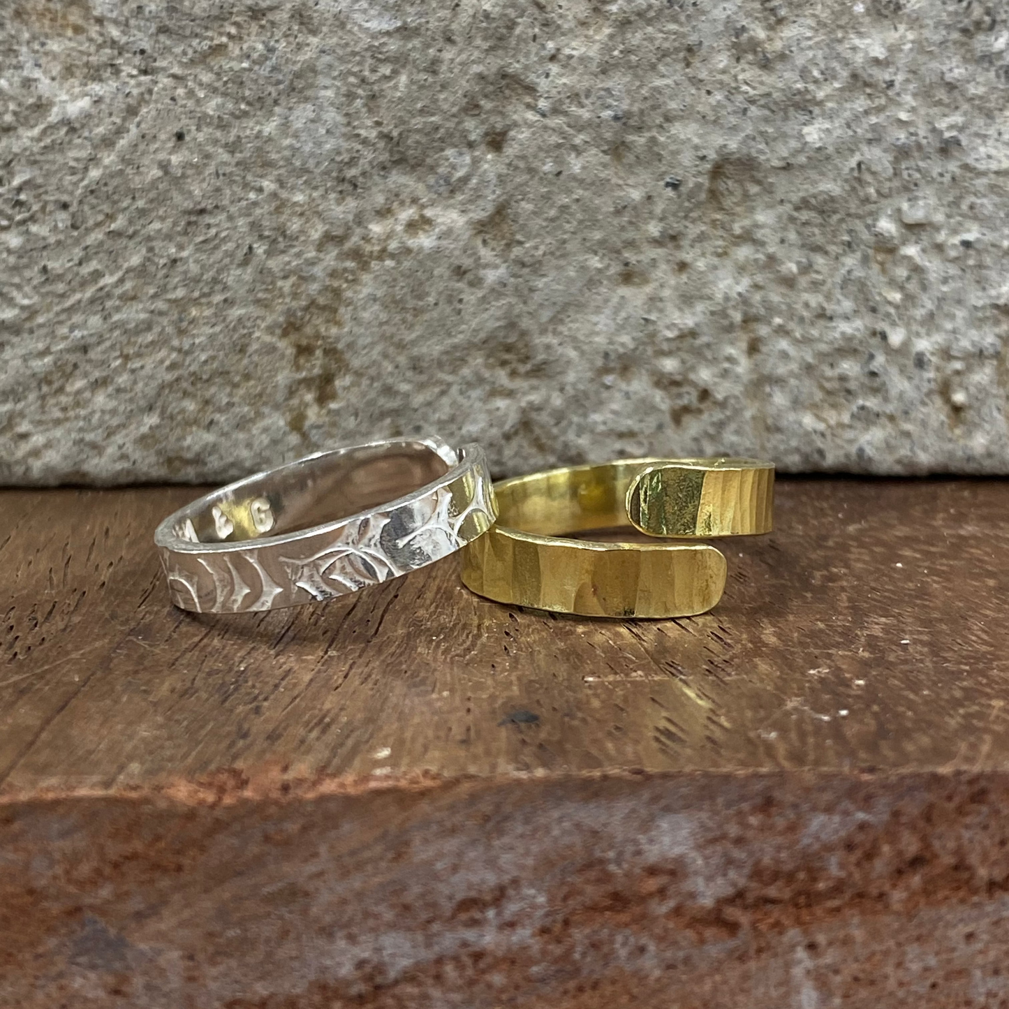 Two open cuff rings, one silver-toned with a hammered leaf pattern and one gold-toned with a textured finish, displayed on a rustic wooden surface against a stone wall.