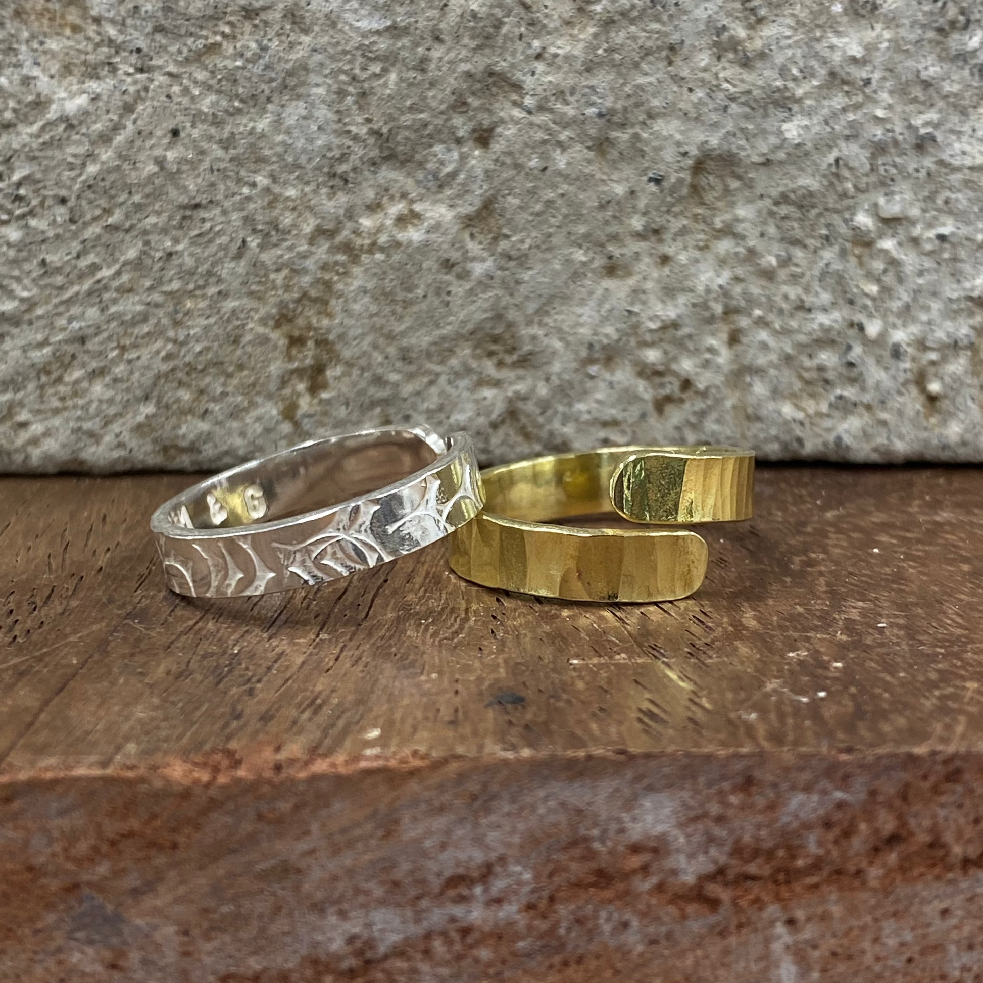 Two open cuff rings, one silver-toned with a hammered leaf pattern and one gold-toned with a textured finish, displayed on a rustic wooden surface against a stone wall.
