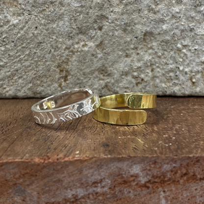 Two open cuff rings, one silver-toned with a hammered leaf pattern and one gold-toned with a textured finish, displayed on a rustic wooden surface against a stone wall.