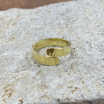 A textured gold open-bend ring resting on a rough, light gray stone surface with a blurred brown wooden background.