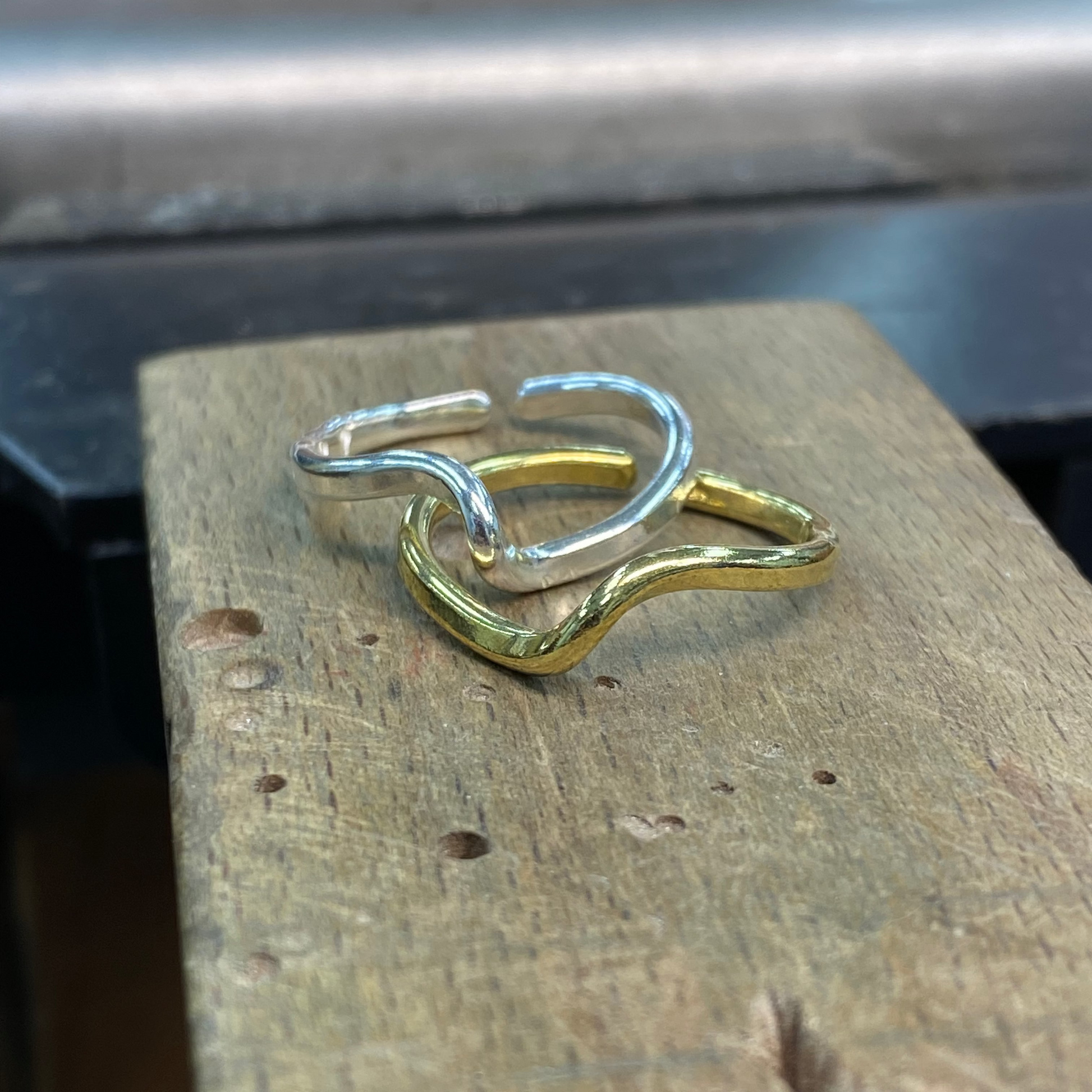 Two wavy open-band rings, one polished silver and one polished yellow gold, interlocked on a rustic wooden block.
