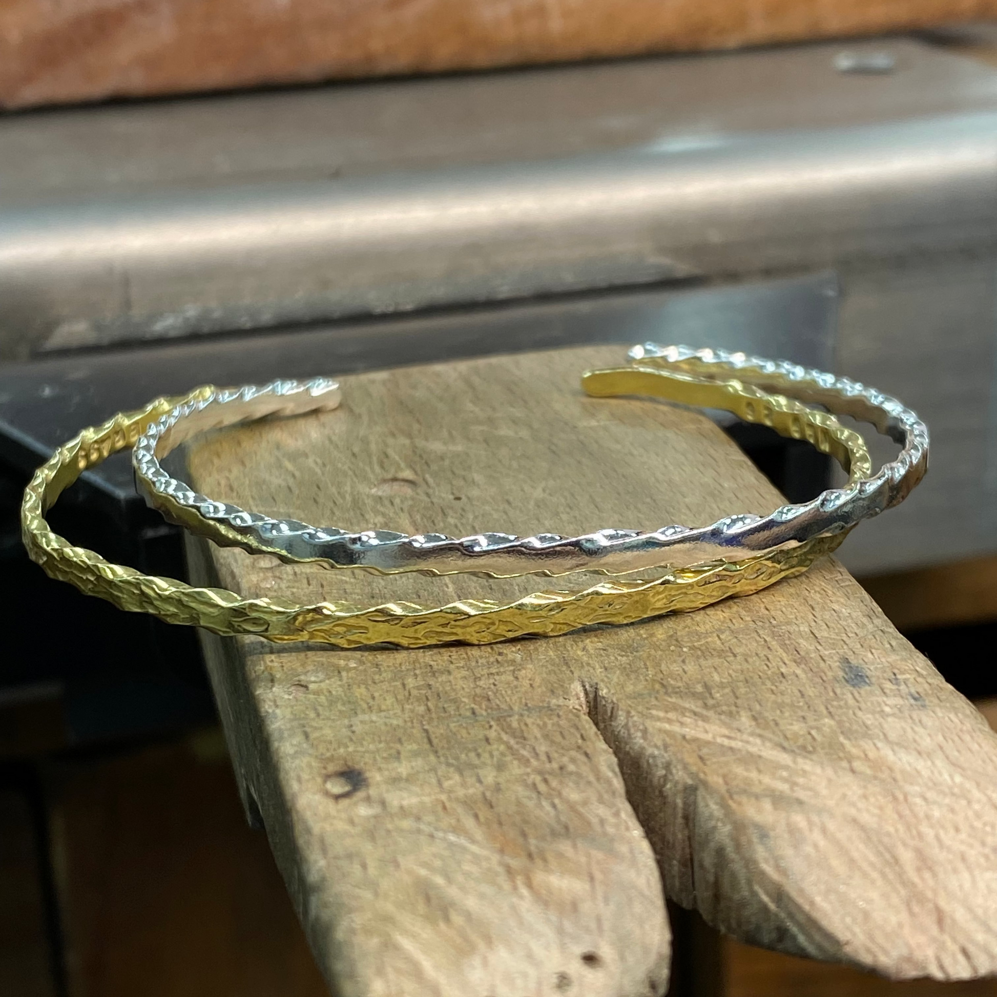 Two textured open cuff bracelets, one in yellow gold and one in silver, displayed on a rustic wooden block.