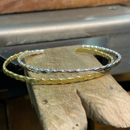 Two textured open cuff bracelets, one in yellow gold and one in silver, displayed on a rustic wooden block.
