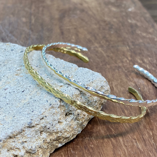 Two textured open cuff bracelets, one gold and one silver, displayed on a light gray stone over a rustic wooden surface.
