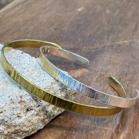 Two hammered metal cuff bracelets, one gold-toned and one silver-toned, displayed on a textured stone slab over a wooden surface.