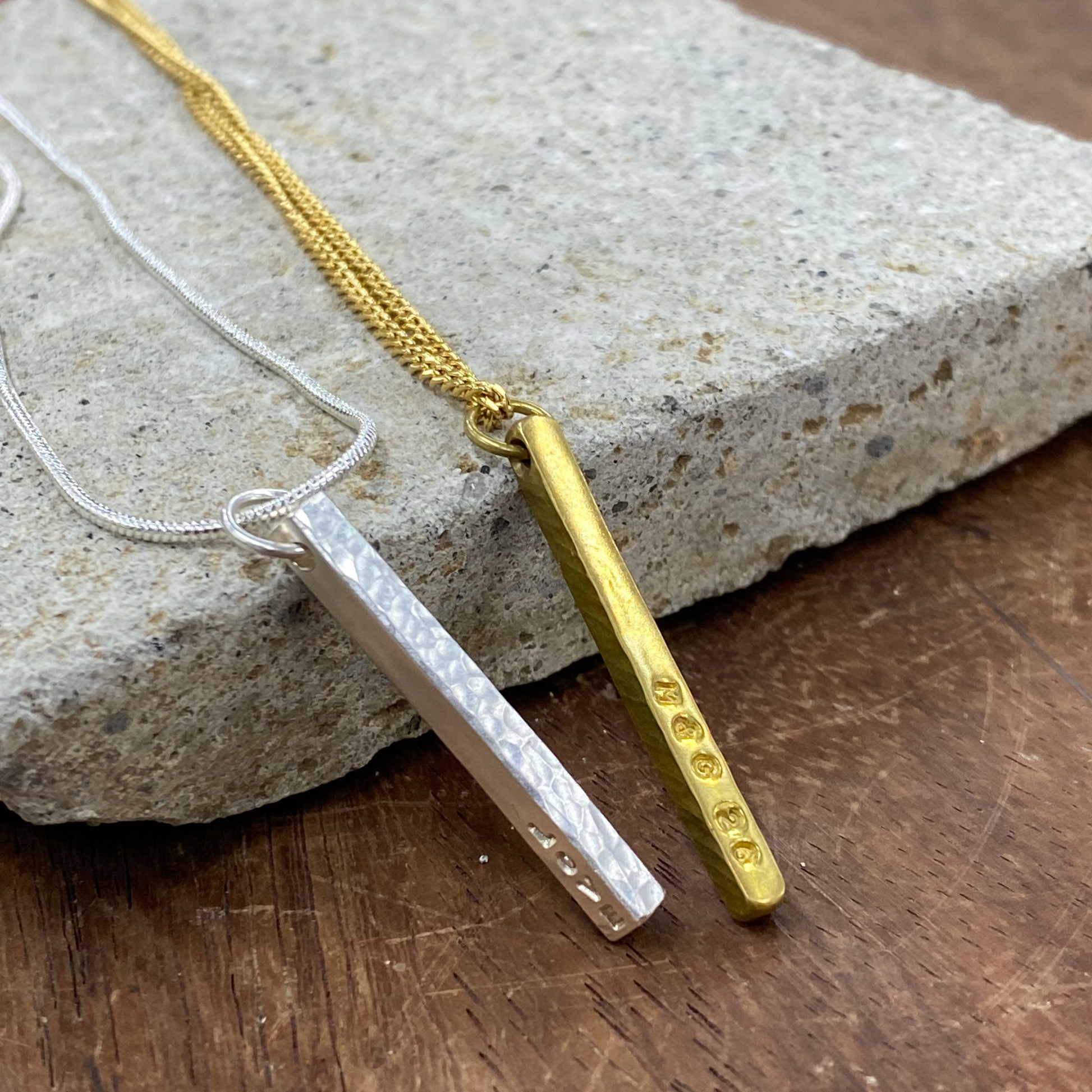 Two bar necklaces displayed on a textured stone slab over a wooden surface. One necklace is gold with a hammered bar pendant, and the other is silver with a hammered bar pendant.