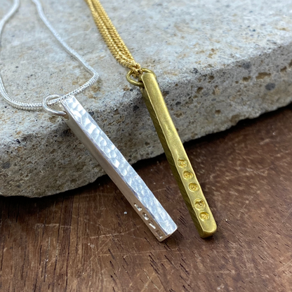 Two minimalist bar necklaces, one silver with a hammered finish and one gold with small engraved dots, displayed on a textured stone over a wooden surface.