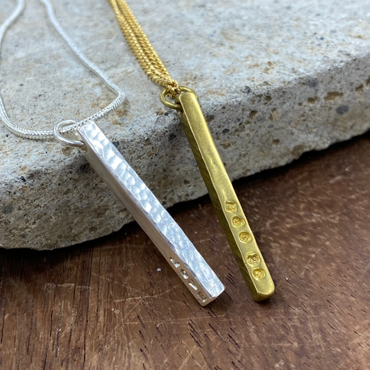 Two minimalist bar necklaces, one silver with a hammered finish and one gold with small engraved dots, displayed on a textured stone over a wooden surface.