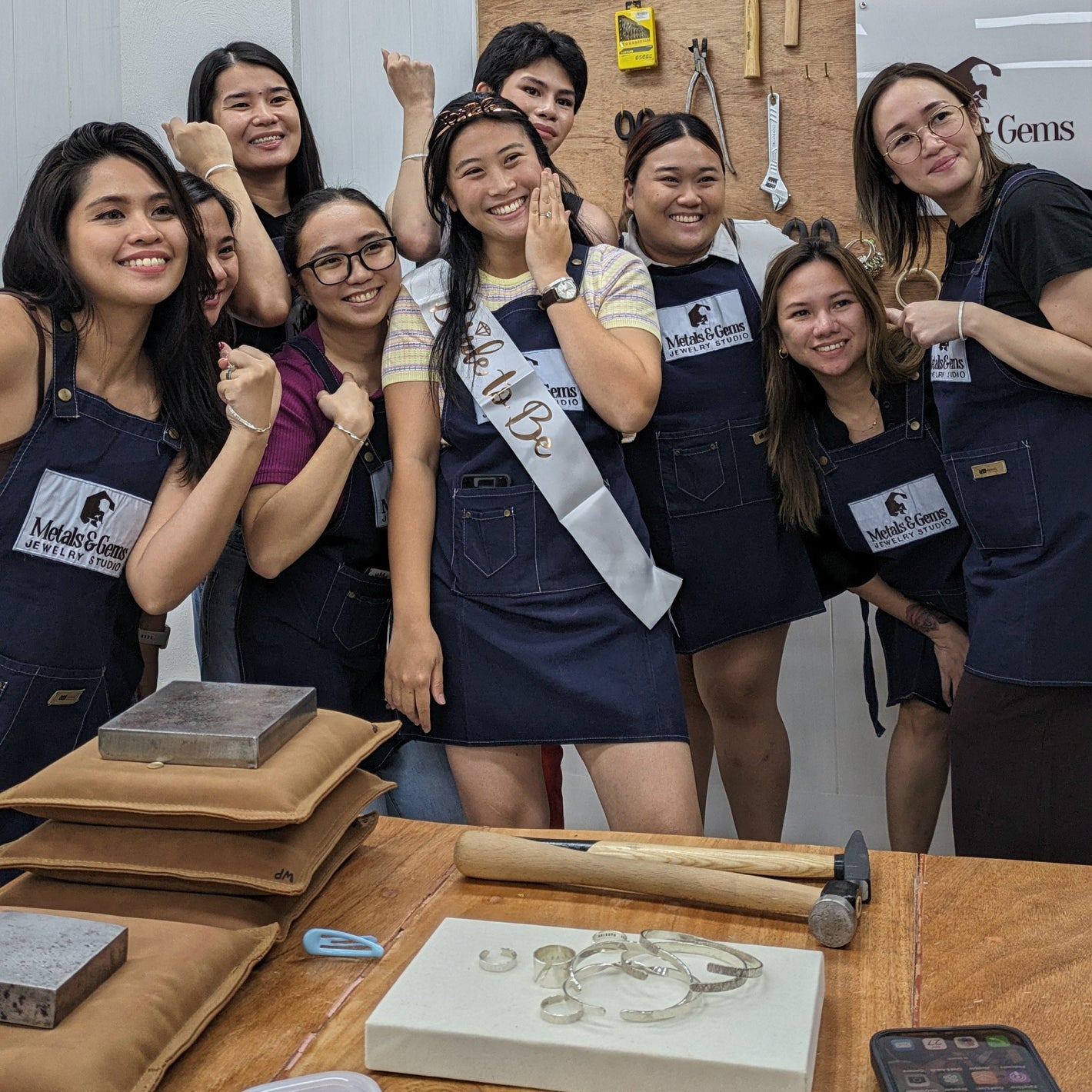 Bridal shower jewelry making group of women posing together in a workshop setting with tools and materials on a table.