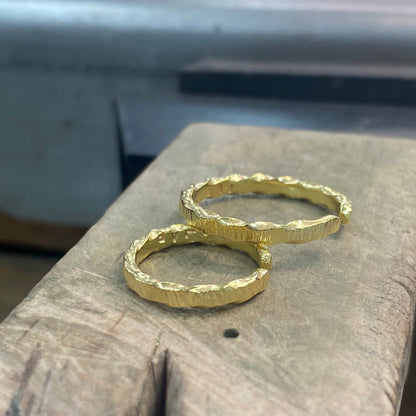 Two textured gold rings resting on a rustic wooden block with a blurred workshop background.