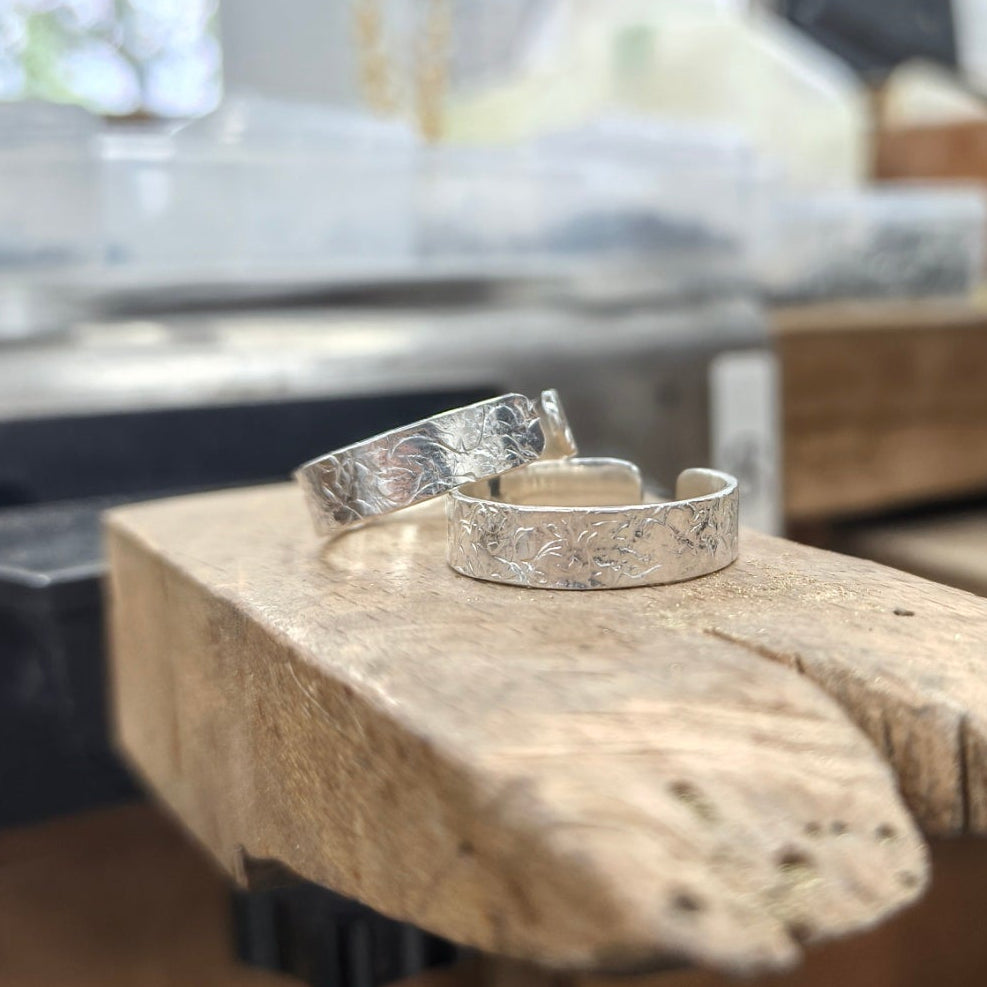 Two textured silver bands, one with a hammered pattern and the other with a carved organic pattern, displayed on a rustic wooden block in a workshop setting.