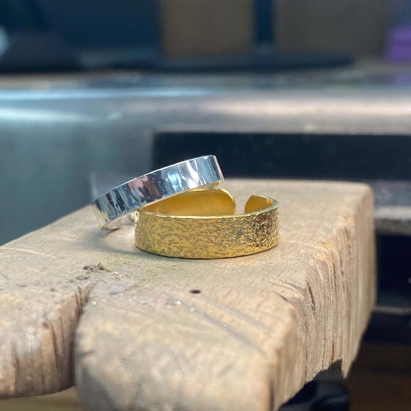 Two textured metal rings, one gold and one silver, displayed on a rustic wooden block with a blurred workshop background.