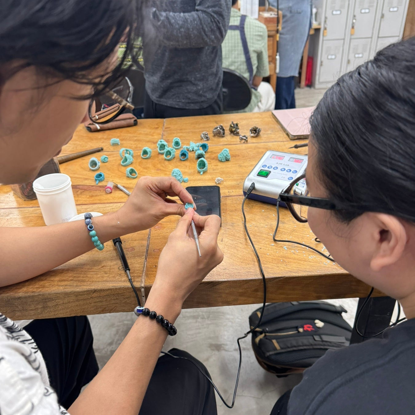 Two people seated at a wooden table in a workshop or studio, working on jewelry-making tools and small turquoise-colored beads. A third person stands in the background near lockers.