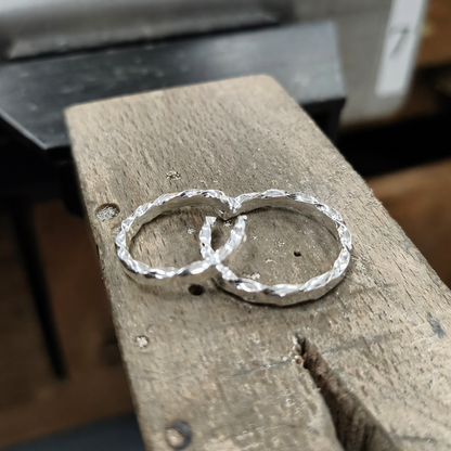 Two textured silver rings resting on a wooden block in a workshop setting.